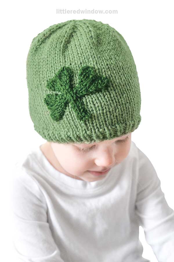 A child wears a charming shamrock hat, featuring a green knit design with a darker green shamrock on the side. Peering down, the child pairs it perfectly with a crisp white shirt.