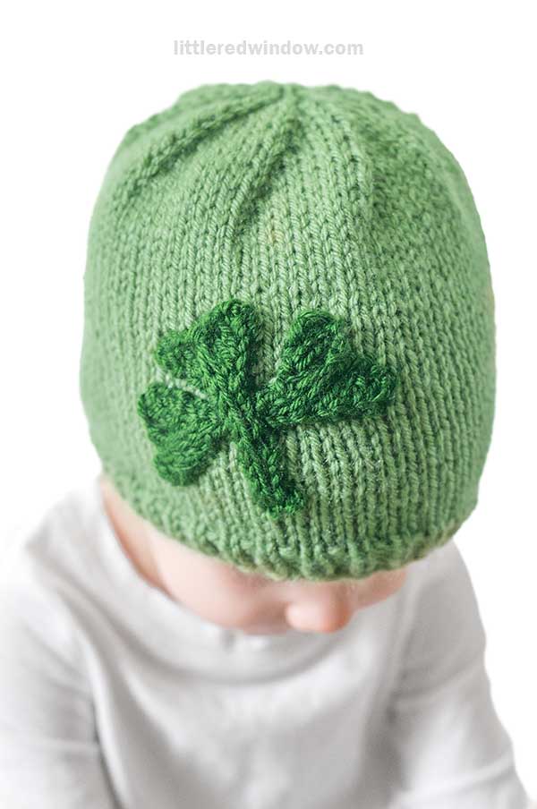 A baby wearing a charming green shamrock hat. The little one is dressed in a light-colored shirt, with eyes gently cast downward.
