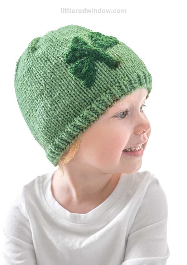 A child wearing a knitted shamrock hat is smiling and looking to the right. The green hat features a large shamrock on the side, perfectly complementing their white shirt against the plain white background.