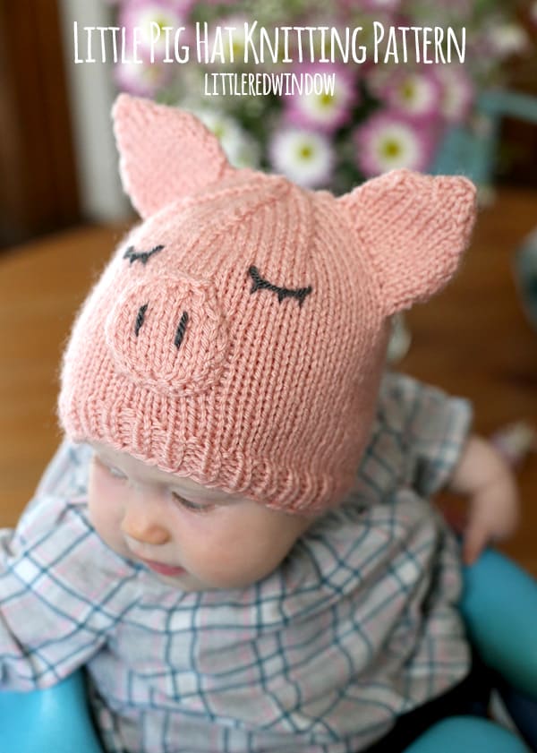 Baby in plaid shirt sitting in a blue chair and wearing a pink knit pig hat looking off to the left
