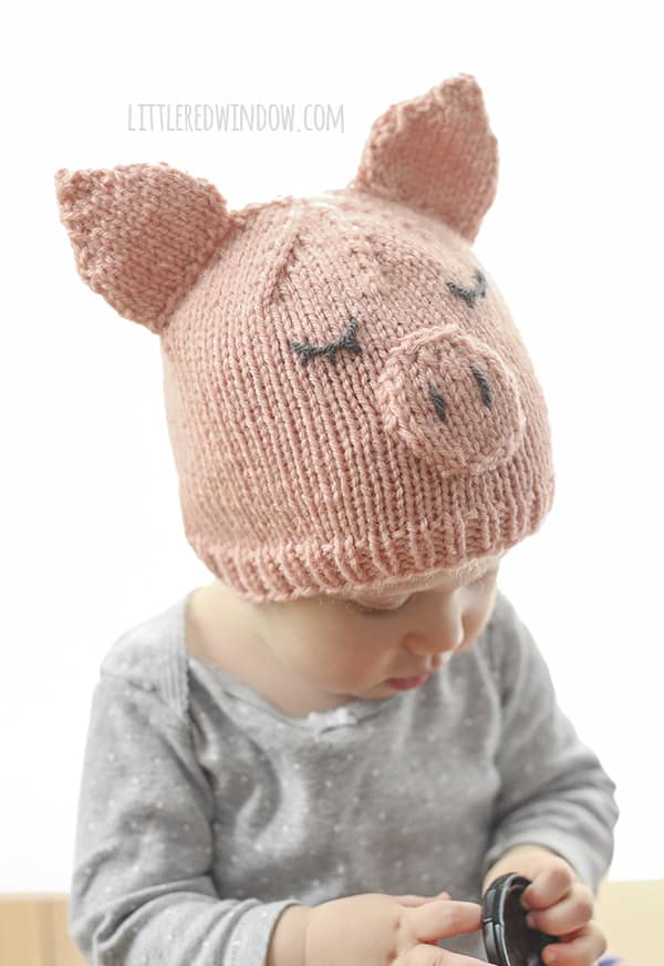 Toddler in a gray shirt wearing a pink knit pig hat and looking at a camera lens cap in their hands