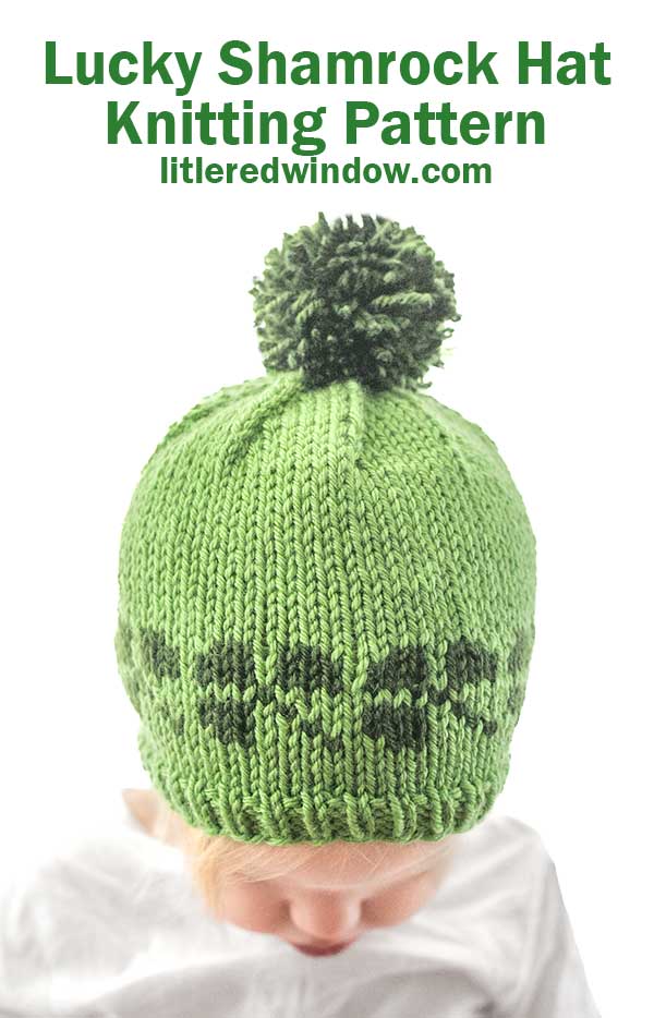 A baby sporting a green knitted Shamrock Hat, adorned with a dark green shamrock pattern and a pom-pom. The text above reads,