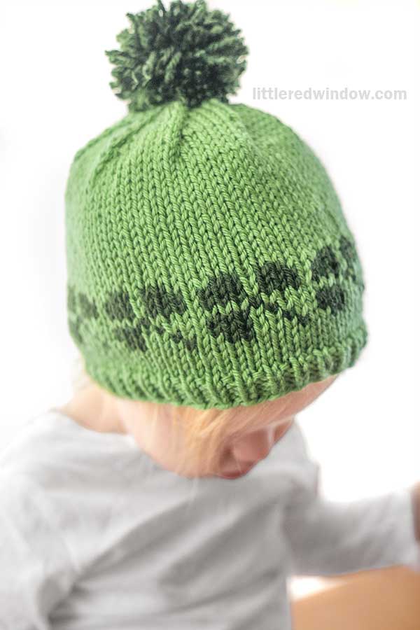 A child wearing a shamrock hat with a green knit design and a pom-pom on top.The child gazes downward against a plain white background.