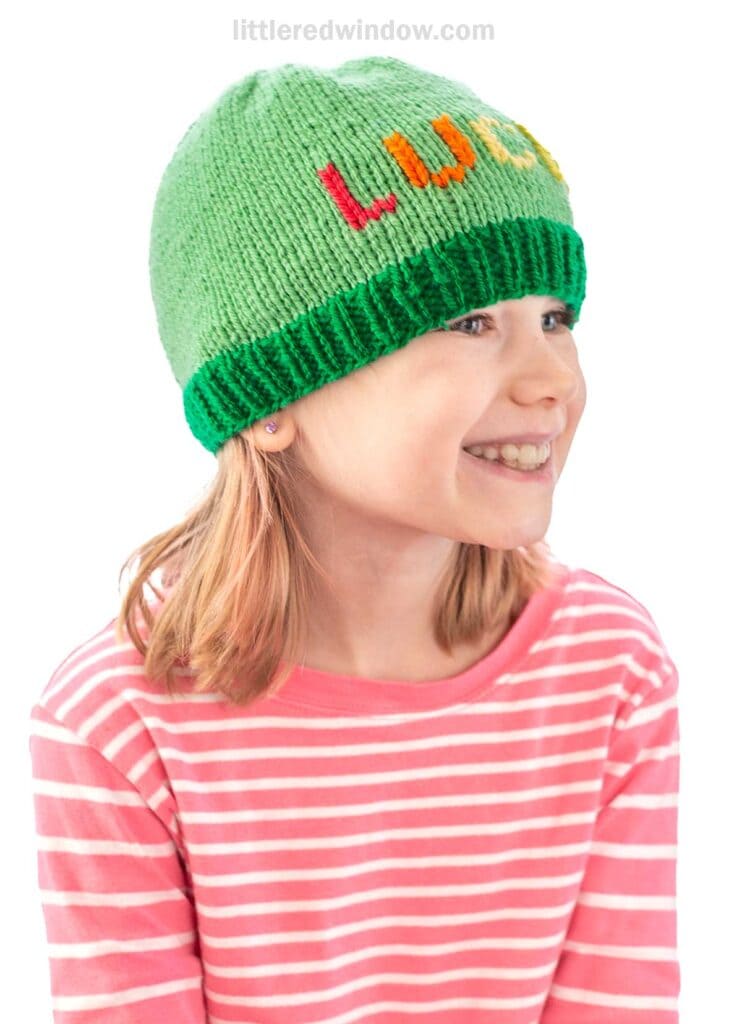 A smiling child wearing a pink and white striped shirt and a green knit hat with colorful rainbow letters that say LUCKY. The background is white.