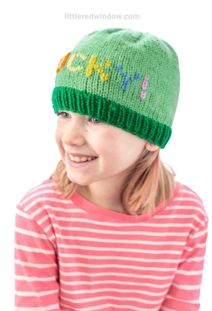 A child with shoulder-length hair smiles while wearing a green knit beanie with the word