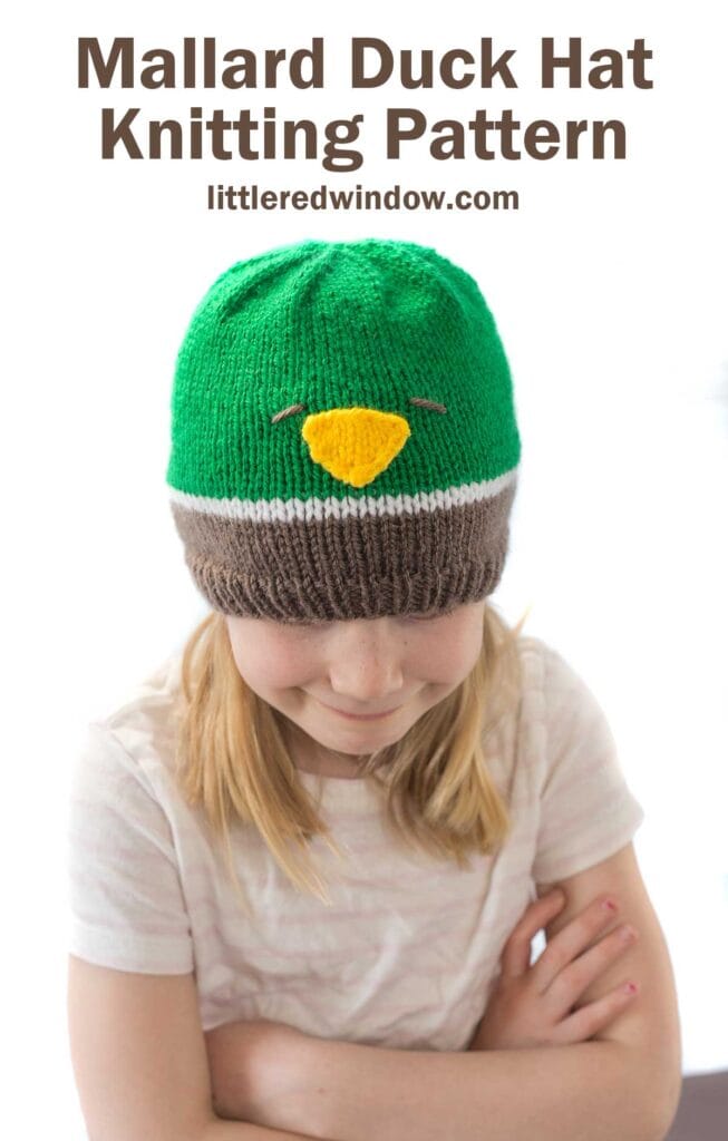 girl wearing a knit hat in brown white and green that looks like a mallard duck looking down at her lap