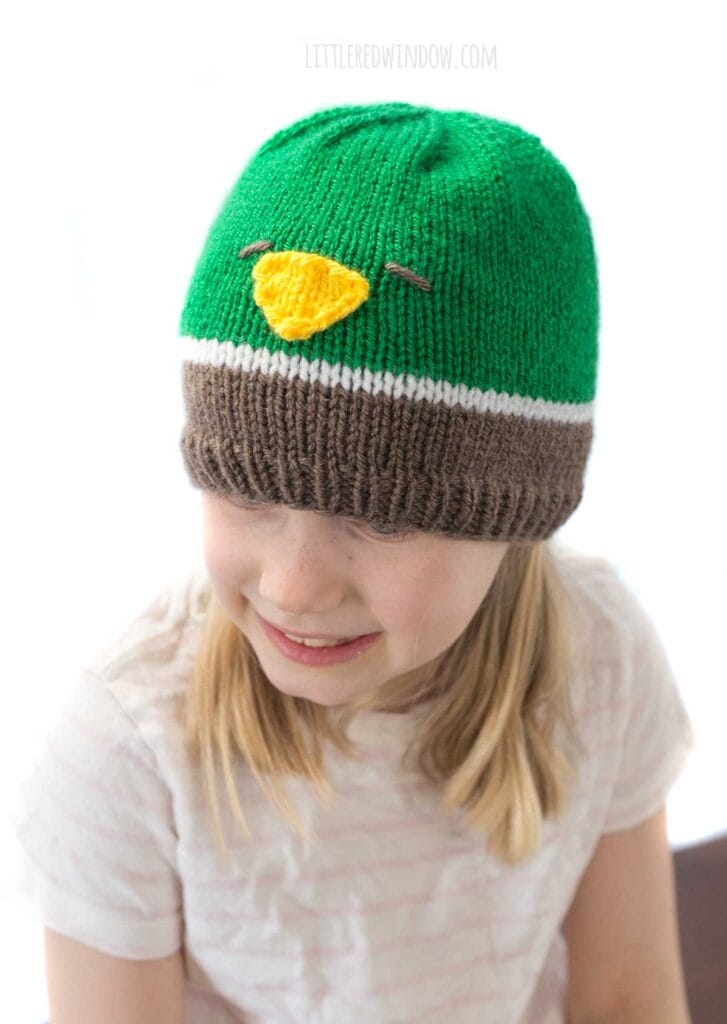 smiling girl in a tshirt wearing a knit hat in brown white and green that looks like a mallard duck looking down and slightly to the left