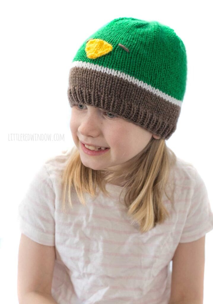 smiling girl in a tshirt wearing a knit hat in brown white and green that looks like a mallard duck looking slightly to the left