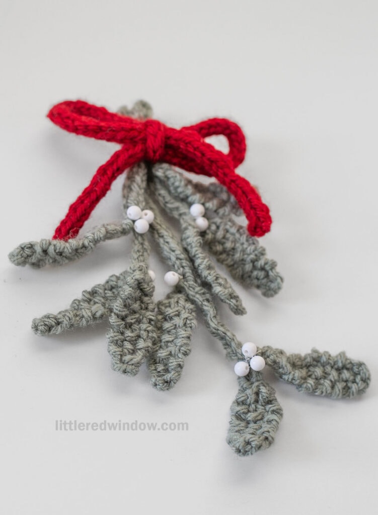 A crocheted sprig of mistletoe with gray-green leaves, white berry clusters, and a red yarn bow on top, displayed on a light surface—perfect inspiration for your next mistletoe knitting pattern.