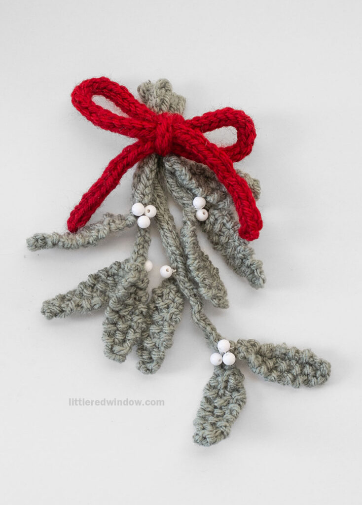 A crocheted sprig of mistletoe with green leaves, white berries, and a red yarn bow on a plain white background—perfect for those seeking inspiration for their next mistletoe knitting pattern.