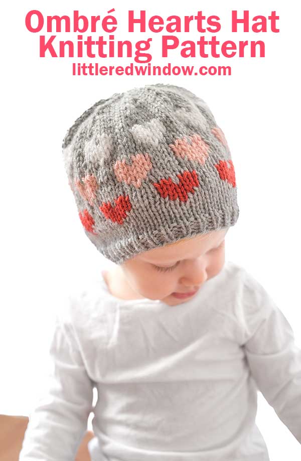 A baby in a gray knitted hat adorned with an ombré heart pattern in pink and red hues looks down, exuding cuteness. The text above says,