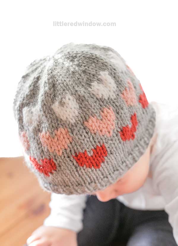 A child wearing a charming heart hat with a knitted pattern of red, pink, and white hearts. The gray beanie frames their face as they look down, showcasing the intricate design that adds a touch of warmth and whimsy.