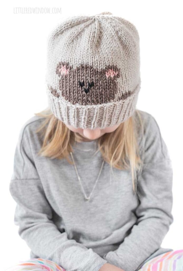 blonde girl in gray shirt looking down wearing a tan knit hat with a folded brim that has a brown teddy bear head peeking over the brim
