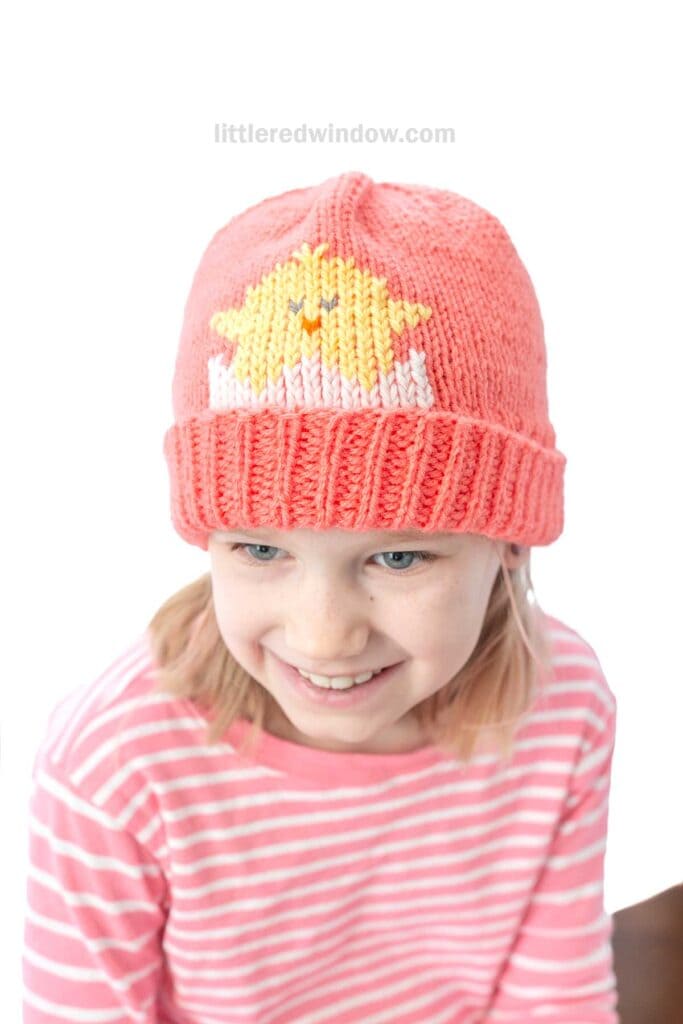 A child beams with joy, sporting a pink knit hat adorned with a yellow star, reminiscent of delightful chick patterns. Paired with a pink and white striped shirt, the ensemble stands out against the plain white backdrop.