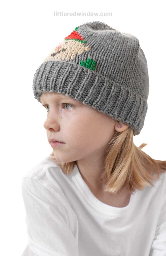 A child wearing a whimsical gray knit beanie with an elf hat pattern sits in a white shirt, looking to the side. The plain white background highlights the child and the beanie.