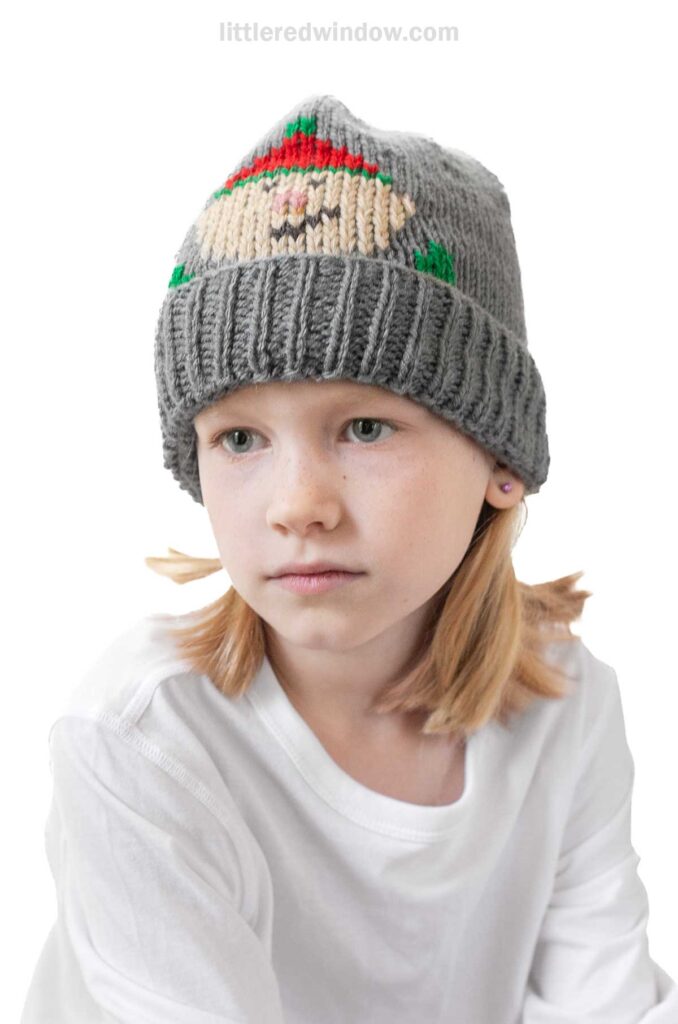 A child in a gray elf hat, crafted with a charming knitting pattern, and a white long-sleeve shirt sits against a white background. The child has shoulder-length hair and a thoughtful expression.