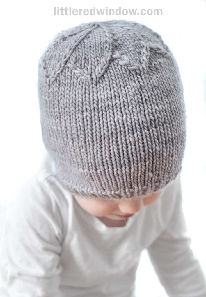 A baby wearing a gray knitted petal hat featuring a leaf design sits in a white long-sleeve shirt against a light background. The baby's face is partially visible, looking down.