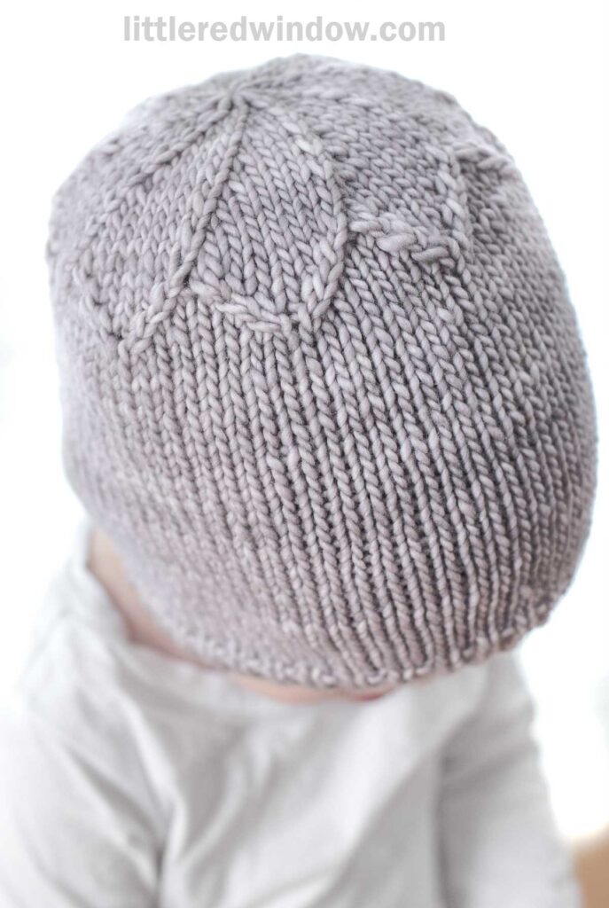 A baby wearing a hand-knitted lavender Petal Hat with a leaf pattern on top is looking downward. The background is blurred, highlighting the texture and detail of the knitted hat. The text