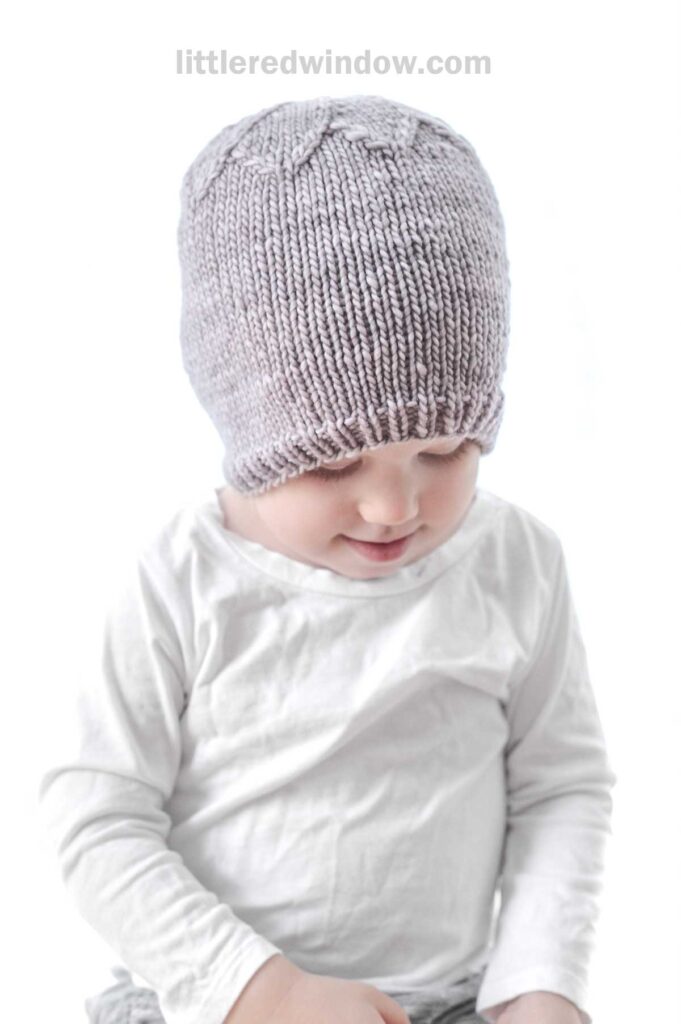 A toddler wearing a light grey knit petal hat and a white long-sleeve shirt looks down with a slight smile. The background is a clean white, highlighting the child and the hat's texture. The website name