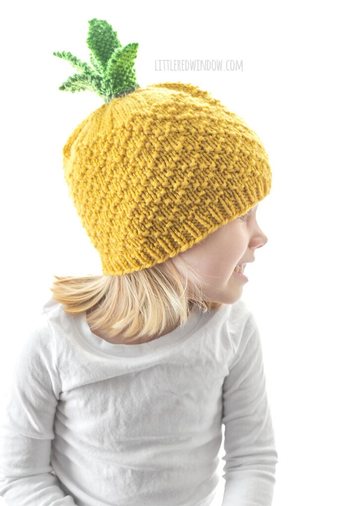 child in white shirt wearing a knit pineapple hat looking off to the right in front of a white background