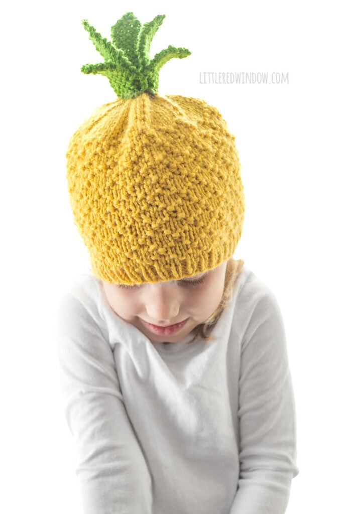 shrugging kid in white shirt wearing a yellow knit pineapple hat with green pineapple leaves on top in front of a white background