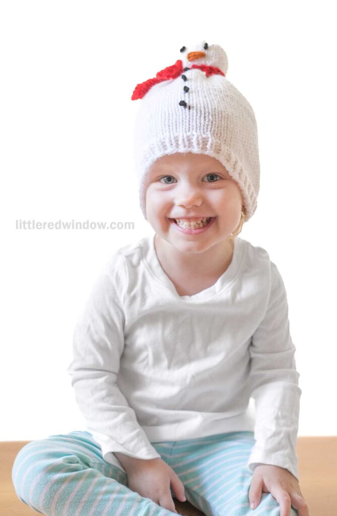 A child sits smiling, donning a snowman hat adorned with small black buttons, an orange nose, and a red scarf. Alongside this charming headpiece, the little one wears a white shirt paired with striped blue and white pants.