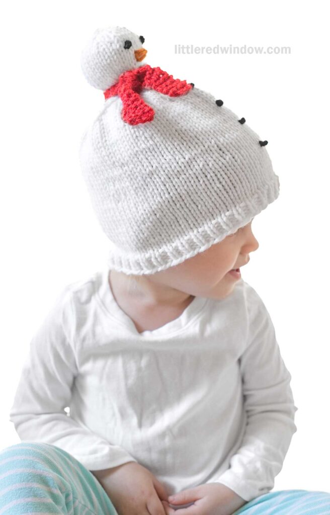 A child dons a whimsical snowman hat, complete with a red scarf and an orange nose, as they sit gazing to the side. The playful snowman design sits prominently atop the cozy white knit.