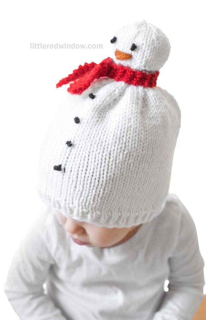A child wears a delightful snowman hat, complete with black button details and a red scarf, perfectly matching the white shirt underneath.