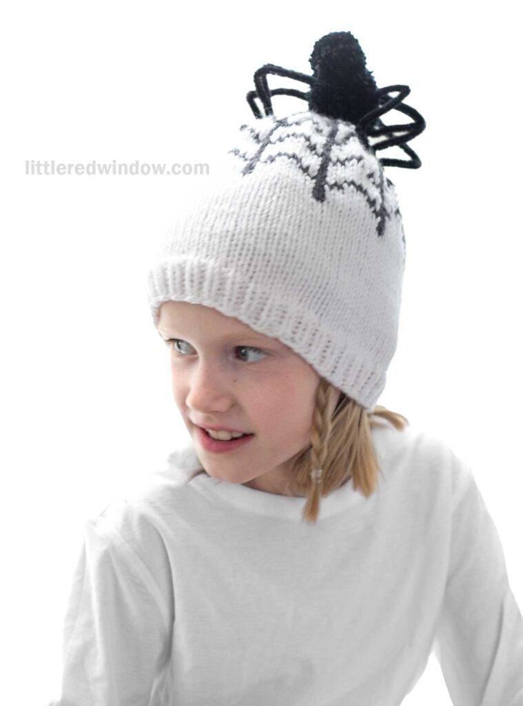 A young child with blonde hair and wearing a white shirt is looking to the side. They are wearing a white knitted hat with a black spider design on top.