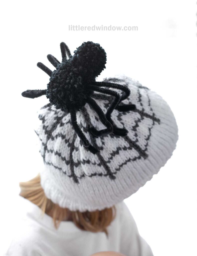A child is wearing a white knit hat with a black spider and spiderweb design. The spider, made of yarn, sits prominently on top of the hat. The child’s face is not visible as they are looking downwards.