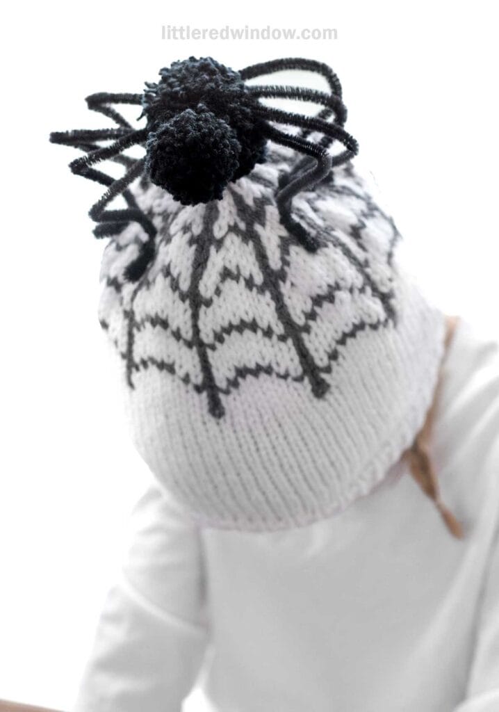 A child wearing a white knit hat with a black spiderweb pattern and a black pom-pom on top with black pipe cleaners resembling spider legs sticking out. The child is dressed in white, and the background is plain white.