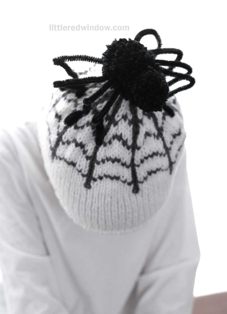 A person wearing a white knitted hat with a black spider web design and a black pom-pom on top. The individual is dressed in a white shirt and is looking downward. The background is white with the text