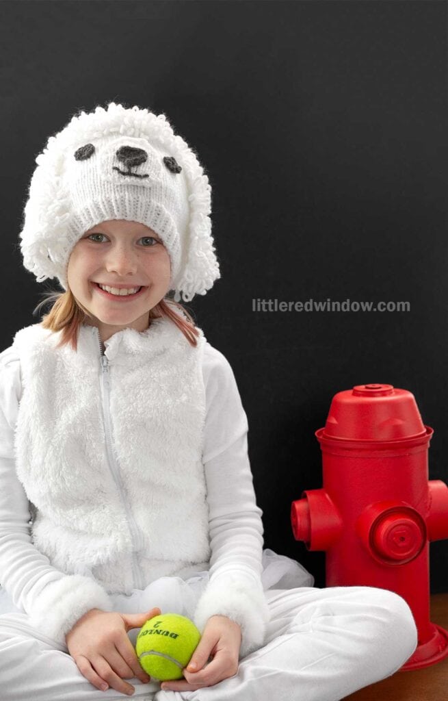A child wearing a fluffy white hat resembling a poodle smiles while sitting next to a red fire hydrant. They are holding a tennis ball. Text reads,