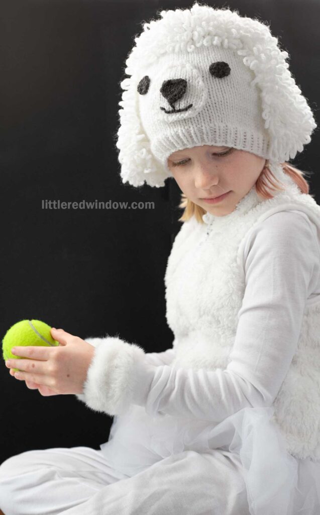 A child wearing a fluffy white poodle outfit and a knitted hat that resembles a white poodle dog's face, holding a green tennis ball. The background is dark, emphasizing the playful costume.