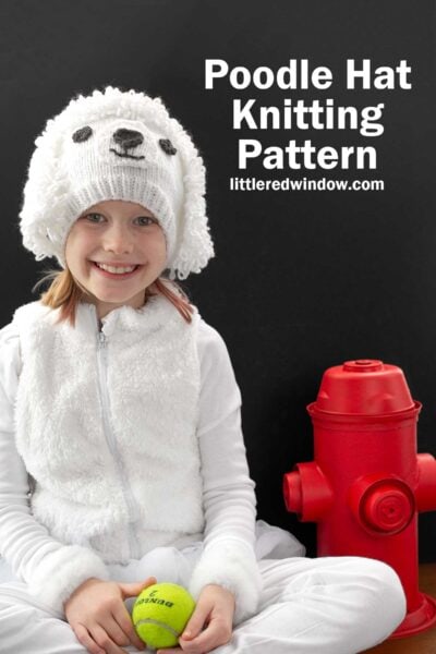 A child wearing a fluffy white hat resembling a poodle smiles while sitting next to a red fire hydrant. They are holding a tennis ball. Text reads, "Poodle Hat Knitting Pattern" and "littleredwindow.com.