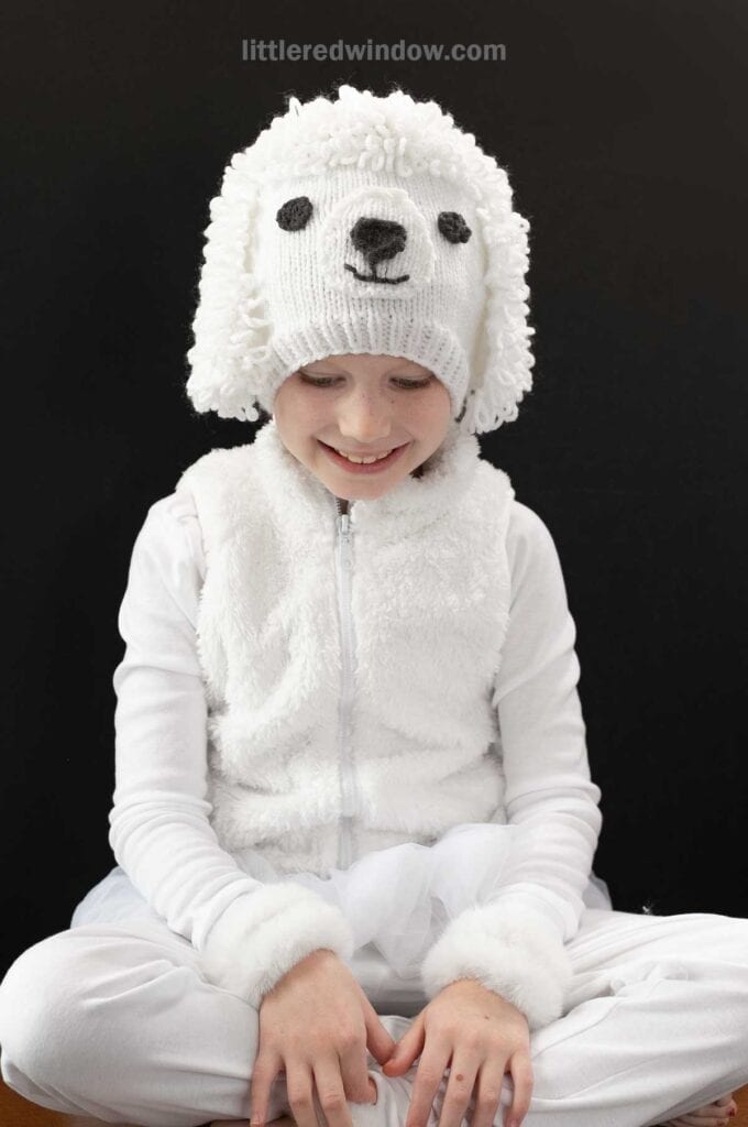 A child wearing a white outfit and a knitted hat resembling a poodle dog's face, complete with floppy poodle ears, is sitting against a dark background. They are looking down and smiling softly.
