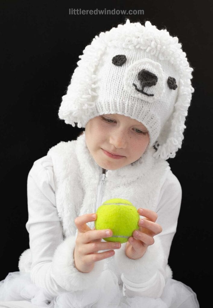 A child wearing a knitted white knit poodle hat and a white outfit holds a yellow tennis ball, looking at it thoughtfully against a black background.