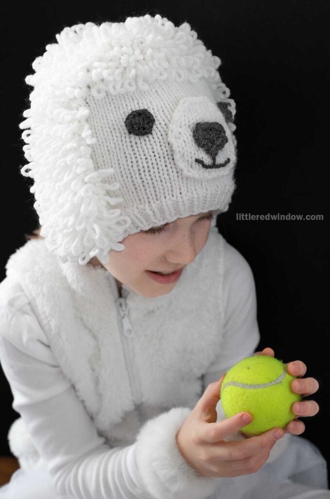 Child wearing a fluffy poodle hat with a knit poodle dog face design, holding a yellow tennis ball against a black background.