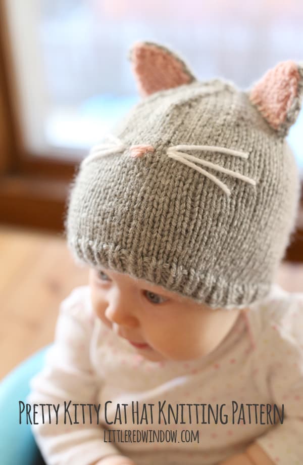 Baby in white onesie sitting in a blue chair wearing a gray knit cat hat with pink and gray hat ears and whiskers on the front looking off to the left