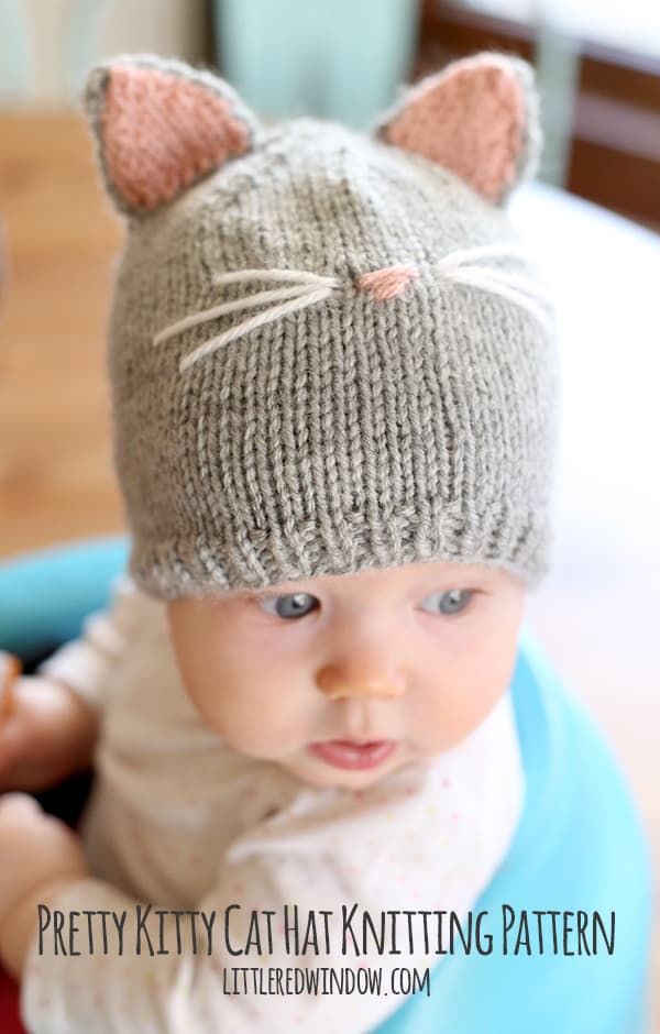 Baby in white onesie sitting in a blue chair wearing a gray knit cat hat with pink and gray hat ears and whiskers on the front looking off to the right