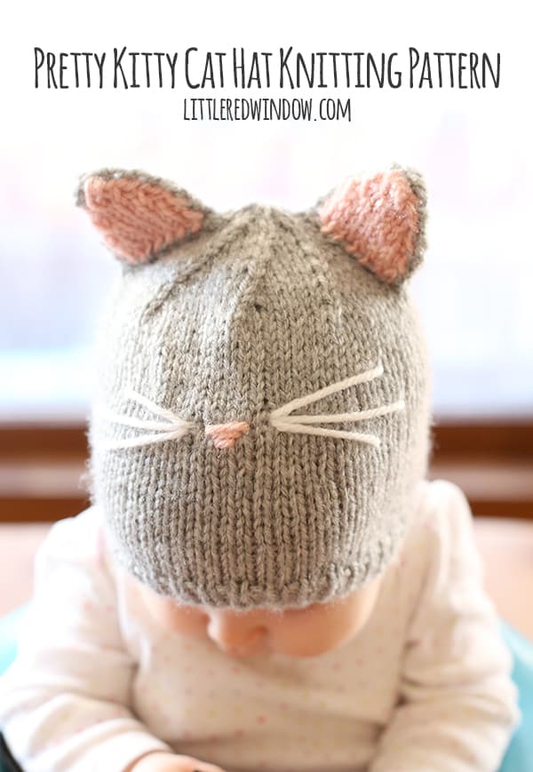 Baby in white onesie sitting in a blue chair wearing a gray knit cat hat with pink and gray hat ears and whiskers on the front