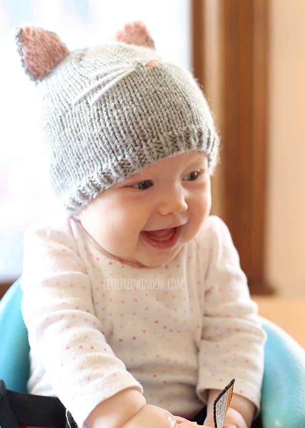 Baby in white onesie sitting in a blue chair wearing a gray knit cat hat with pink and gray hat ears and whiskers on the front laughing and looking to the right