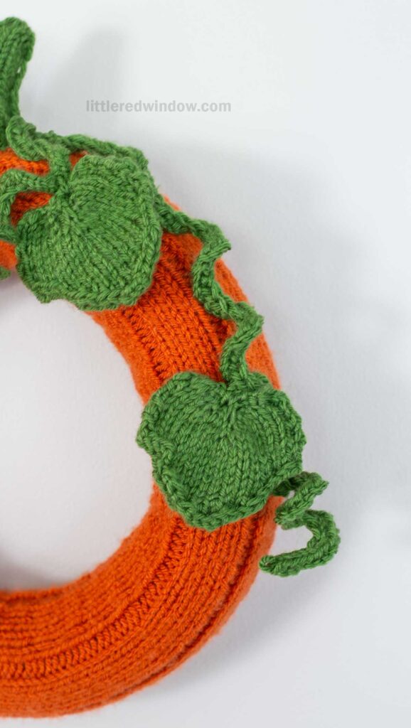 A close-up of a pumpkin wreath, featuring an orange knitted ring decorated with green knitted leaves and curly vines, displayed against a plain white background.