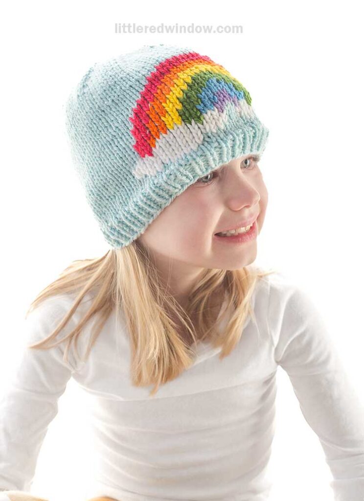 A child beams joyfully, wearing a light blue knitted hat showcasing a vibrant rainbow, crafted from a delightful knitting pattern. Paired with their white shirt, the ensemble exudes cheerful charm.