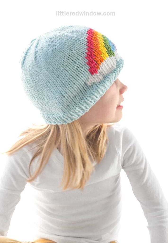 A child in a light blue knit hat adorned with a rainbow pattern glances to the side, dressed in a crisp white long-sleeve shirt. The backdrop is brightly lit, highlighting the intricate knitting pattern of the charming headwear.