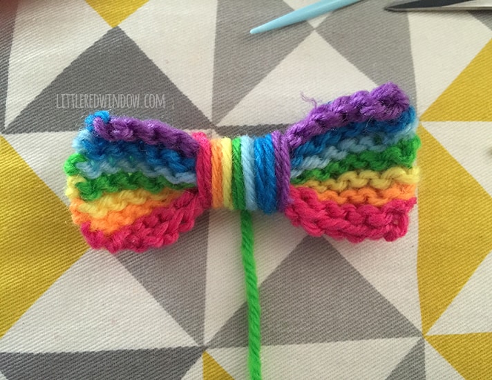 rainbow striped knit bow with rainbow pieces of string wound around the center and one green string hanging down to use to sew the bow to the hat
