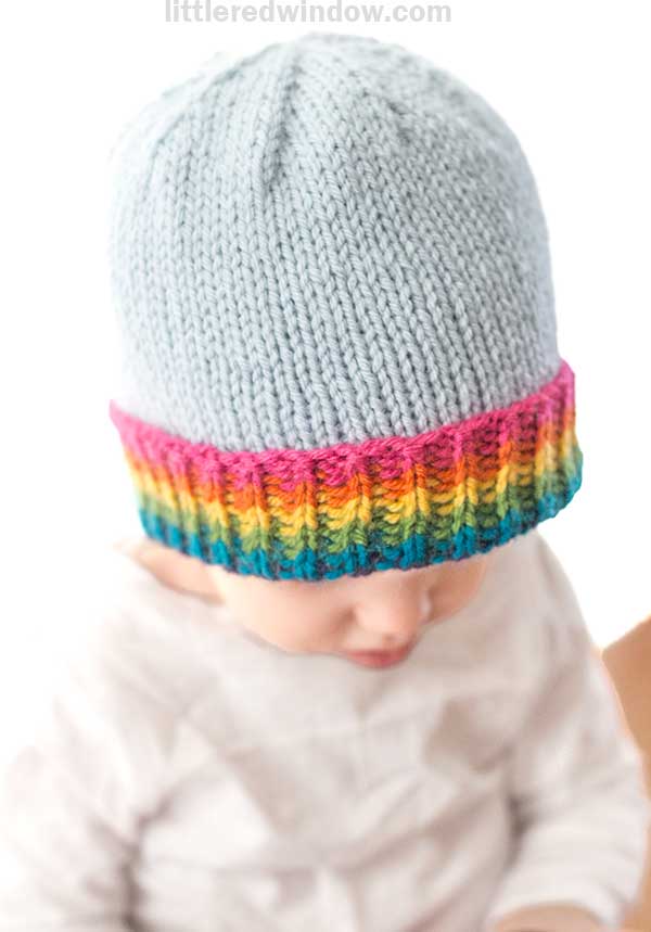 A baby wearing a light gray knitted hat with a vibrant rainbow brim tilts their head slightly down, perfectly complementing their light-colored shirt.