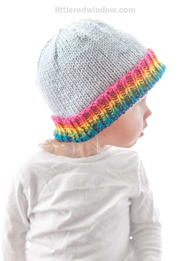 A child in a white knitted hat with a vibrant rainbow brim gazes off to the side, dressed in a simple white shirt. The backdrop is pure white, highlighting the playful touch of color on the hat's brim.