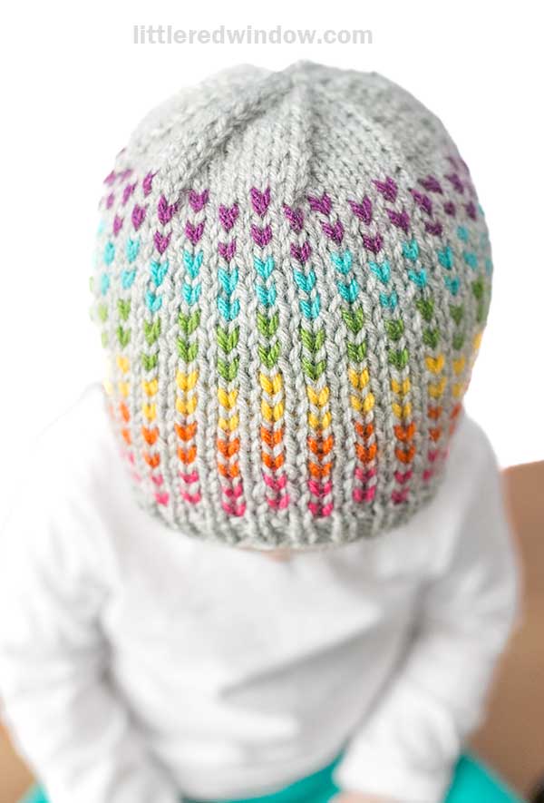 A child wearing a gray knit hat adorned with rainbow dots and colorful vertical streaks of purple, teal, green, yellow, and orange sits facing downward. The child is dressed in a white shirt against a plain background.