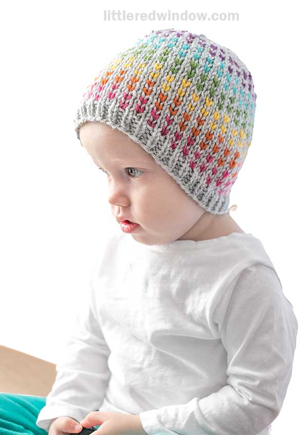 A baby wearing a colorful, knit hat sits facing left. The hat features a rainbow gradient pattern sprinkled with rainbow dots and a gray brim. The baby is dressed in a plain white shirt against a simple background.